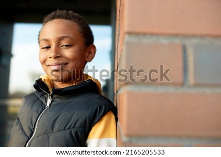 Outdoor picture of handsome confident happy boy of african ethnicity standing over red brick wall, looking at camera with cunning smile dressed in yellow hoodie and black puffer vest