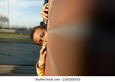 Outdoor picture of little adorable african american boy of 9-years-old playing hide and seek hiding behind red brick wall, showing forehead and eyes. Selective focus. Happy childhood