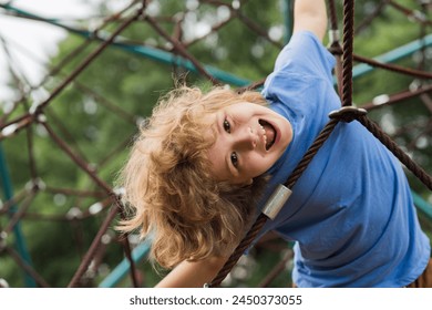 The outdoor playground for children in summer park. Kid play on playground under the tree. Portrait of excited blonde kid doing rock climbing with greenery in the background. - Powered by Shutterstock