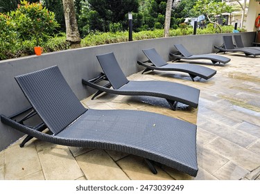 Outdoor poolside lounge chairs or pool side chair with modern black rattan design set against lush greenery, providing a serene relaxation area on wet, clean pavement at a tropical resort. - Powered by Shutterstock