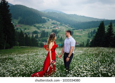 Outdoor portrait of the attractive smiling couple holding hands on the daisy meadow at the background of the beautiful green mountains. The girl is holding the bouquet of wild flowers. - Powered by Shutterstock