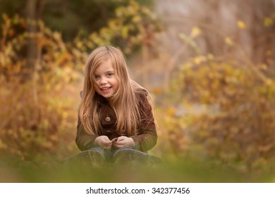 Outdoor Portrait In Autumn Of A Laughing Beautiful Little Girl With Long Hair Sitting On A Grass, Short Depth Of Field