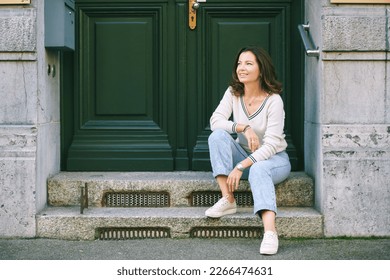 Outdoor portrait of beautiful 40 year old woman sitting on step next to green door, old money fashion style - Shutterstock ID 2266474631