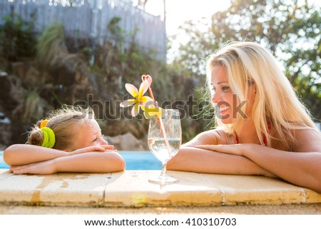 Outdoor portrait of beautiful blonde mother and her cute daughter. Little girl look to her mammy at the pool. Big glass with water, straw and frangipani stay on the pool edge. Happy Mothers day.