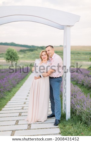 Outdoor portrait of beautiful cheerful romantic mature couple, man hugging woman, standing under the wooden arch in lavender field