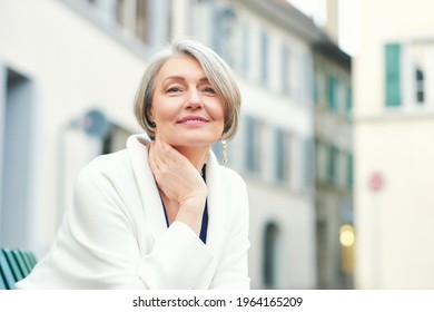 Outdoor Portrait Of Beautiful And Elegant Middle Age 55 - 60 Year Old Woman, Wearing White Jacket, Sitting On Bench Outside