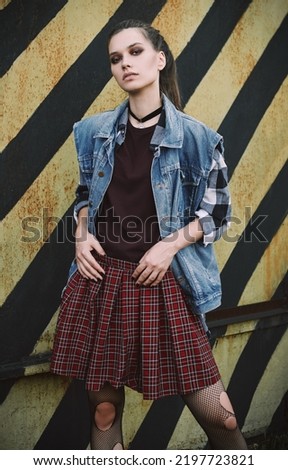 Outdoor portrait of beautiful grunge (rock) girl standing at the wall. Informal model dressed in jean jacket, checkered skirt, shirt and holey tights