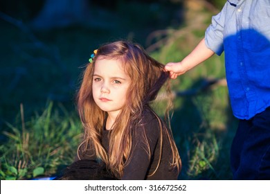 Outdoor Portrait Of A Beautiful Little Girl While Her Toddler Brother Is Pulling Her Hair