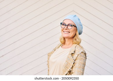 Outdoor portrait of beautiful middle age woman, posing next to white wooden wall, wearing glasses and blue hat