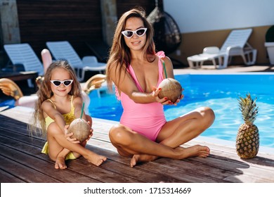 Outdoor Portrait Of Beautiful Woman Mother In Bikini And Her Cute Daughter In Swimsuit. Little Girl And Her Mom Look To The Camera And Smile. Stretch In The Hands Of Coconut.Happy Family At The Pool.