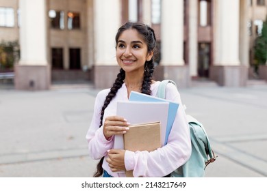 Outdoor Portrait Of Cheerful Indian Female Student With Backpack And Workbooks Standing Near College Building, Looking At Camera And Smiling