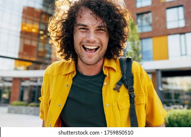 Outdoor Portrait Of Cheerful Man With Curly Hair, Smiling Broadly, Posing For Social Advertisement In The City Street. Happy Student Hipster Male Has Joyful Expression While Walking Outside.