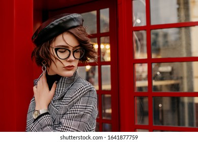 Outdoor Portrait Of Confident Fashionable Woman Wearing Classic Checkered Coat, Wrist Watch, Eyeglasses, Black Faux Leather Beret, Posing Near Red Phone Booth. Copy, Empty Space For Text