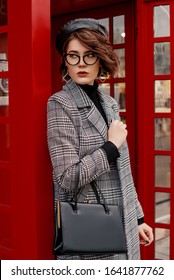 Outdoor Portrait Of Confident Fashionable Woman Wearing Classic Checkered Coat, Glasses, Beret, Holding Classic Leather Handbag, Posing Near Red Phone Booth