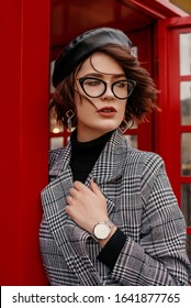 Outdoor Portrait Of Confident Fashionable Woman Wearing Classic Checkered Coat, Wrist Watch, Eyeglasses, Black Faux Leather Beret, Posing Near Red Phone Booth