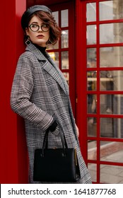 Outdoor Portrait Of Confident Fashionable Woman Wearing Classic Checkered Coat, Glasses, Beret, Holding Classic Leather Handbag, Posing Near Red Phone Booth