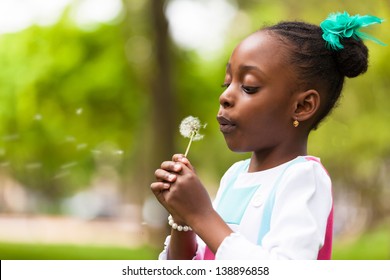 Outdoor Portrait Of A Cute Young Black Girl Blowing A Dandelion Flower - African People