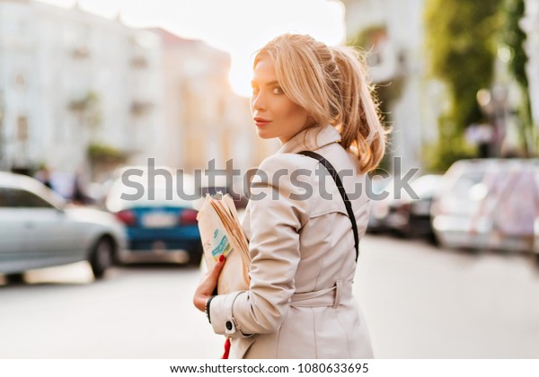 Outdoor\
portrait of elegant yong woman carrying newspapers after work and\
posing with serious face expression. Cute girl in trendy coat looks\
back standing on the street in sunny\
day.