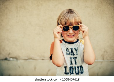 Outdoor Portrait Of Funny Little Boy Wearing Sunglasses And T-shirt With Sign 