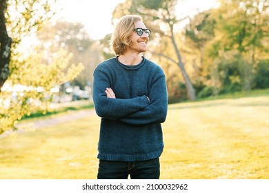 Outdoor Portrait Of Handsome 35 - 40 Year Old Man With Red Hair, Posing In Green Sunny Park, Wearing Blue Pullover And Sunglasses, Arms Crosses