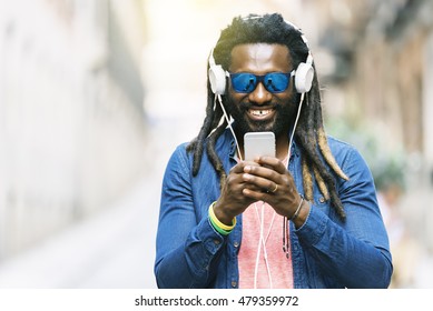 Outdoor Portrait Of Handsome African Young Man Writing Message And Listening Music.