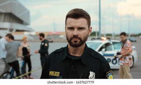 Outdoor Portrait Of Handsome American Cop In Sunglasses Looking Serious And Confident Staying In The Street In Crime Investigation Area With Police Car In Background.