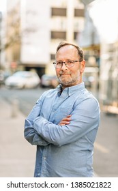 Outdoor portrait of handsome middle age man wearing long sleeve Oxford shirt, posing on city street, arms crossed