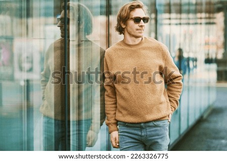 Outdoor portrait of handsome young man wearing sunglasses and beige fuzzy fleece sweater, posing next to mirror wall on city street background
