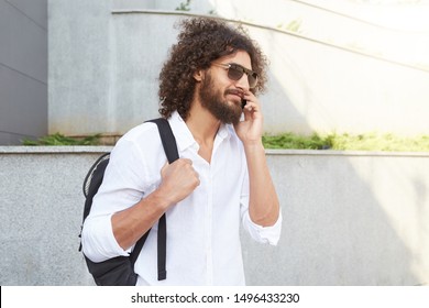 Outdoor portrait of hansome young man with curly hair and lush beard walking down the street while talking to phone, wearing white shirt and black backpack - Powered by Shutterstock