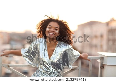 Outdoor portrait of happy glamour african american woman with glitter on face wearing festive wear sequin dress standing on roof terrace during party or celebration and smiling happily at camera