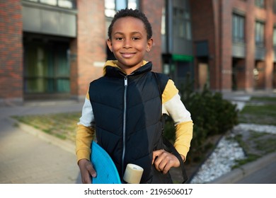 Outdoor Portrait Of Happy Handsome Confident African American Boy Kid Holding Blue Skateboard, Looking At Camera Putting Hand On Waist, Going To School Or Skate Park. Kids Urban Activity