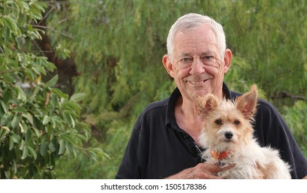 Outdoor Portrait Of A Happy, Healthy 80- Year Old Man Holding His Dog, A Wire Haired Terrier Mix