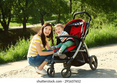 Outdoor Portrait Of A Happy Mother And Son. Baby Sitting In Stroller And Mom Walk In The Summer Park