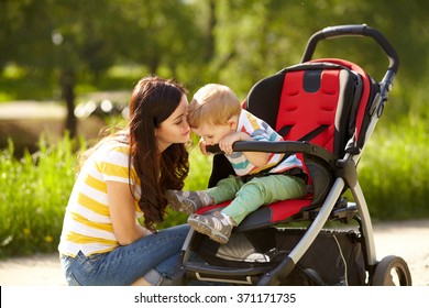 Outdoor Portrait Of A Happy Mother And Son. Baby Sitting In Stroller And Mom Walking In The Summer Park