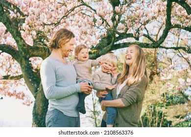 Outdoor portrait of happy young family playing in spring park under blooming magnolia tree, lovely couple with two little children having fun in sunny garden