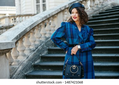Outdoor portrait of pretty curly lady wearing trendy autumn checkered dress, leather beret, wrist watch, holding small black bag, posing in street of European city. Copy, empty space for text