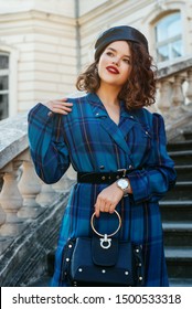 Outdoor portrait of pretty curly lady wearing trendy autumn blue tartan dress, black wide suede belt, leather beret, wrist watch, holding small black bag, posing in street of European city