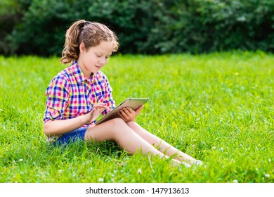 Outdoor portrait of a pretty teenager girl in casual clothes sitting on the grass with digital tablet on her knees, reading and surfing