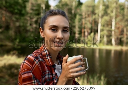 Outdoor portrait of pretty woman tourist in red checkered shirt holding mug of tea on background with river and forest, camping on lake, enjoying morning in wild nature, looking at camera