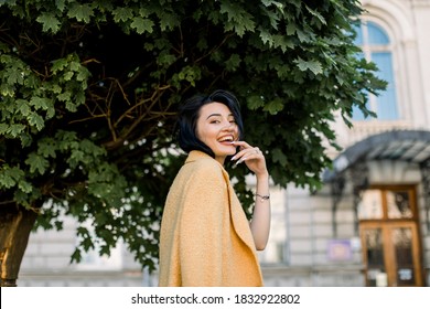 Outdoor portrait of pretty young mixed raced laughing girl in yellow blazer, posing to camera and enjoying her city walk. Elegant Asian woman sitting on bench with city building and tree on background