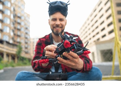 Outdoor portrait of smiling casually dressed bearded man setting generic design fpv multicopter drone in his hands while sitting at the street. Focus is on hands with drone.