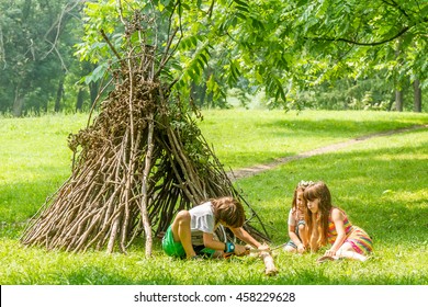 Outdoor Portrait Of Three Happy Kids - Boy And Girls - Playing Next To Wooden Stick Hut House, Looking Like Indian Tepee, On Natural Background