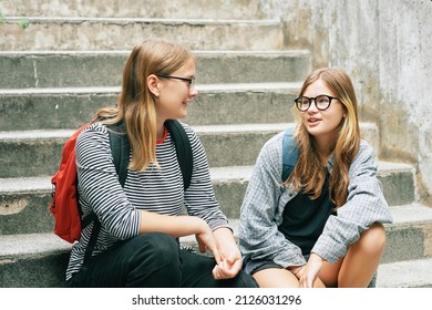 Outdoor portrait of two teenage girl sitting on stairs, wearing backbacks, talking to each other