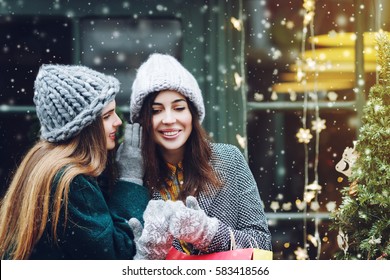 Outdoor Portrait Of Two Young Beautiful Fashionable Girls Posing With Colorful Shopping Bags. Woman Whispering To Her Smiling Friend. Ladies Wearing Stylish Winter Hats. Christmas Background, Snowfall