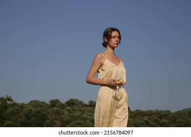 Outdoor Portrait Of Woman In Beige Silk Outfit Holding A Glass Of White Wine. Idyllic Summer Vacation Concept.	