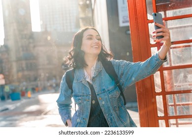 Outdoor portrait of woman using a mobile phone and taking selfie  against a red phonebox in the city of England - Shutterstock ID 2236133599