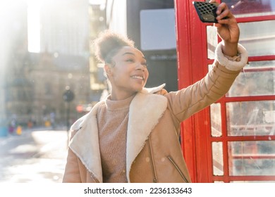 Outdoor portrait of woman using a mobile phone and taking selfie  against a red phonebox in the city of England - Shutterstock ID 2236133643