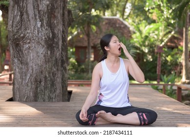 Outdoor Portrait Of Young Asian Woman Sitting Under Big Tree, Being Bored And Sleepy, Yawning While Practicing Yoga. Laziness Concept.