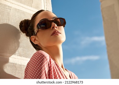 Outdoor portrait of a young beautiful confident woman posing on the street. Model wearing stylish sunglasses. Girl looking up. Female fashion. Sunny day. Close up. City lifestyle. Copy space for text