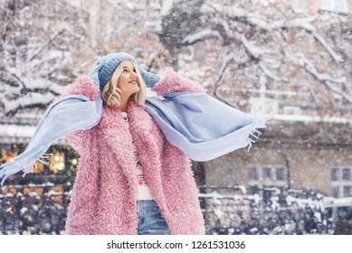 Outdoor portrait of young beautiful fashionable happy smiling girl wearing trendy pink faux fur winter coat, light blue beanie hat, scarf, posing in snow covered street. Copy, empty space for text